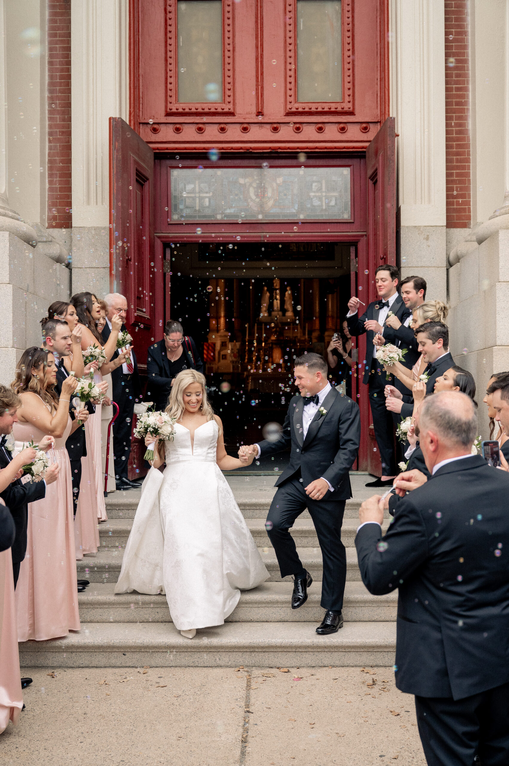 Bride and groom walk out of the Church of the Gesu following their wedding ceremony. Guests blow bubbles and celebrate.